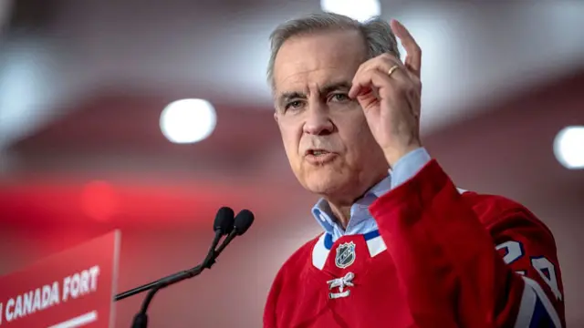 Carney speaks into microphones while wearing a red hockey shirt