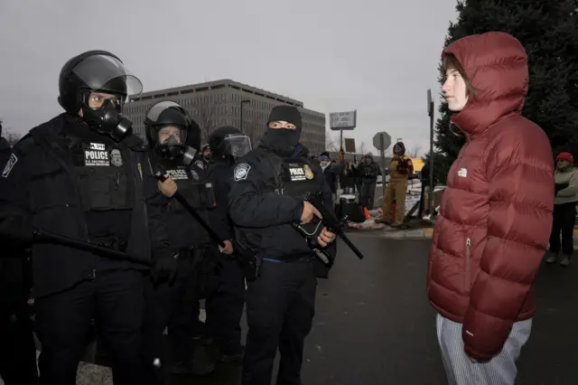 A demonstrator wearing a red puffer jacket and striped baggy trousers stands in front of U.S. Customs and Border Protection (CBP) agents holding batons and smoke masks during a protest outside the Whipple Federal Building