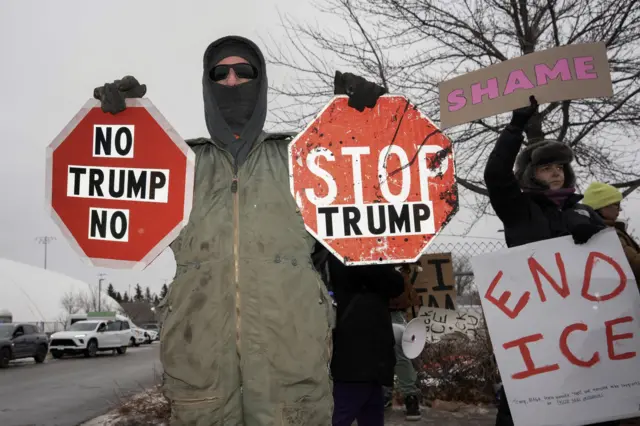 A protestor is wearing a khaki jumpsuit, a dark hoodie and balaclava and sunglasses and two stop signs that say "Stop Trump" and "no Trump no"