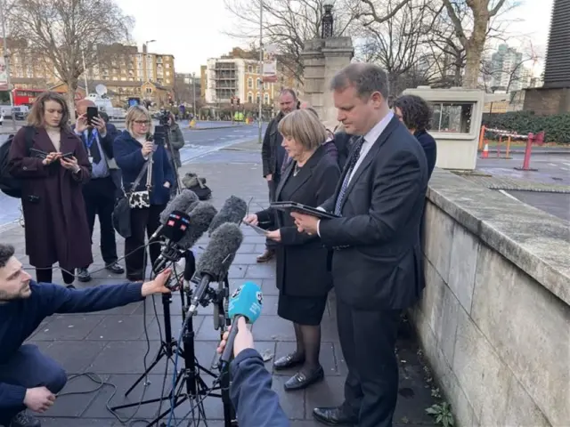 A man and a woman outside Inner London Crown Court. They are reading from tablets and are surrounded by journalists.