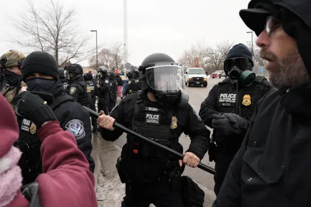 Three ICE agents stand with batons and gas masks. In the foreground are two civilians who might be protestors, one with their fist raised. One ICE agent has his baton clutched as if he may swing it