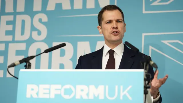 Robert Jenrick speaks from a Reform UK podium at a press conference in London.