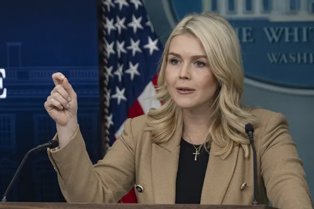 White House press secretary Karoline Leavitt wearing a tan blazer, black top underneath and a necklace with a cross on it. Her hand is in a fist with her thumb and index finger together and there is an American flag behind her.