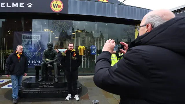 Watford fans pose next to the Graham Taylor statue at Vicarage Road