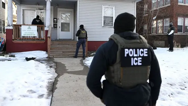 ICE agents, wearing dark coloured clothes and vests that read "Police ICE", stand around a house. There is snow on the ground outside