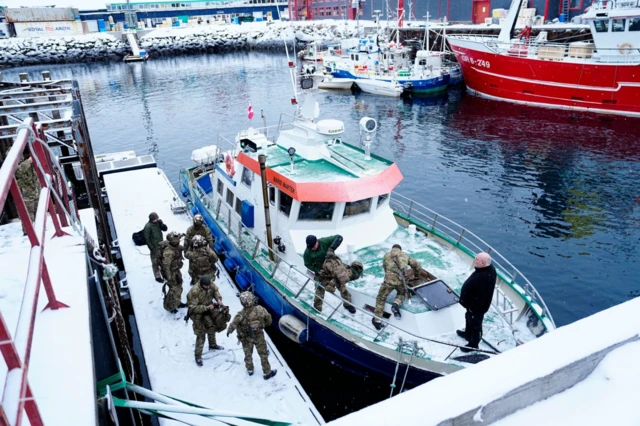 Wide shot of a berth at the port of Nuuk. Six Danish soldiers in military fatigues have disembarked, two remain on a small vessel moving equipment