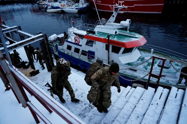 A group of Danish soldiers in military fatigues disembark a small boat at Nuuk port. Two of them carrying equipment up a set of snow-covered wooden steps