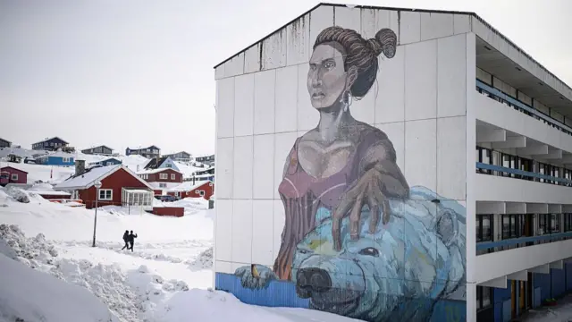 A residential building in Greenland with a large mural of a woman stroking a polar bear, which is lying down. The ground is covered in snow and two people are walking past