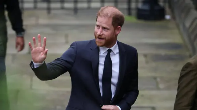 Prince Harry waving outside of court. He's wearing a dark suit and tie with a white shirt.