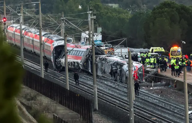 Wide image shows a train with first two carriages derailed and emergency workers next to it, in Adamuz