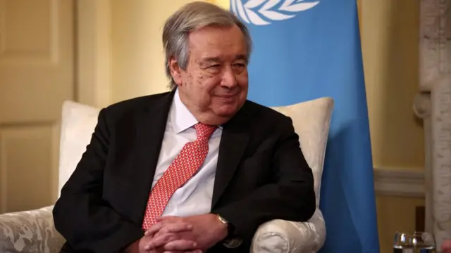 Guterres sits with hands clasped in front of UN flag