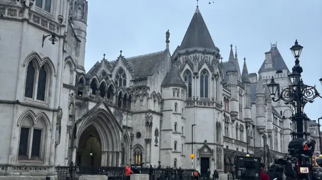 An old court building in overcast London morning. There's some camera crews poised outside. The building is grand with gothic arches and lots of small towers.