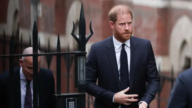 Prince Harry walking outside the court building. He's wearing a dark suit and looks very serious