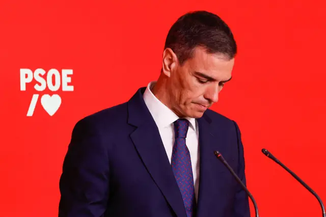 Close up shot of Pedro Sánchez looking down as he speaks into a microphone at an event. He is wearing a dark suit, white shirt, and purple purple and red tie.