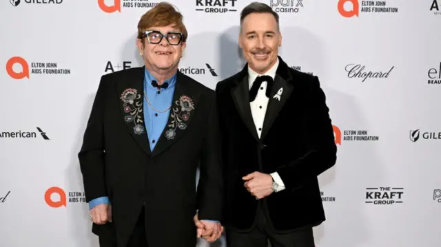 Elton John and David Furnish at a press conference. They are both wearing black suits - Elton John is has flowers embroidered onto his lapels and is wearing a blue shirt. David Furnish is wearing a tuxedo with a loose bow tie. They are holding hands.