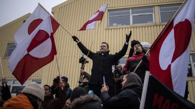 Greenland's premier Jens-Frederik Nielsen waving a Greenland flag and spreading his arms wide, amid around a dozen other protestors waving flags