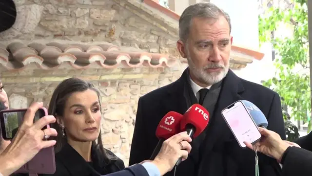 The Spanish Queen and King standing next to each other and speaking to the media, with several microphones in front of them