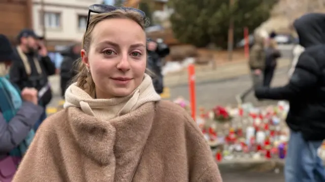 A woman wearing a brown coat with sunglasses on her head. A vigil can be seen blurred behind her.