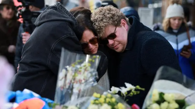 Three people hug each other by floral tributes at Le Constellation bar