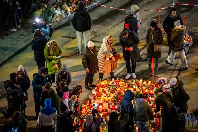 A makeshift memorial is made out of candles near the bar, there are many people gathered around it, many wearing thick winter clothes