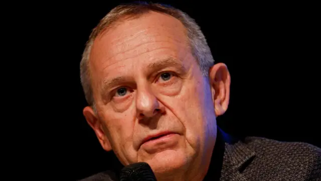 Eric Bonvin speaking at a news conference. He is holding a microphone and wearing a dark jacket and shirt