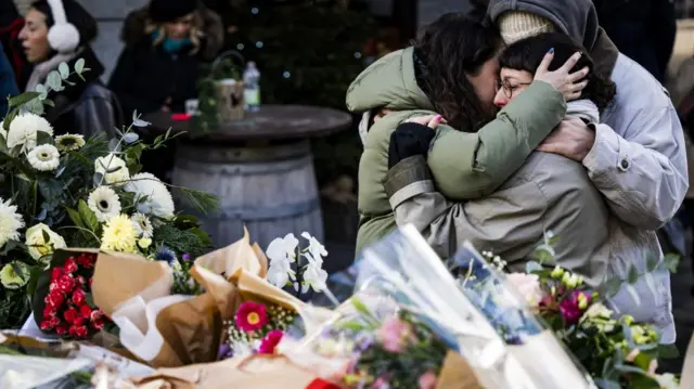 Three women hug each other in front of a floral tribute