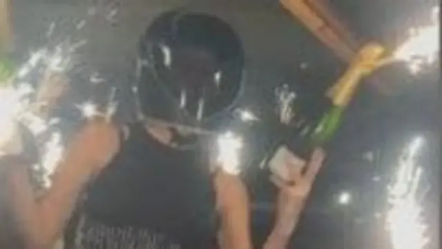 A person in a crash helmet holding a bottle of champagne with a sparkler attached