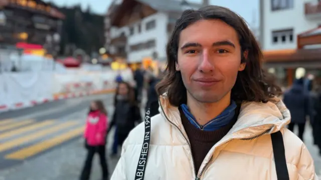 A boy with shoulder length brown hair wearing a white puffer jacket