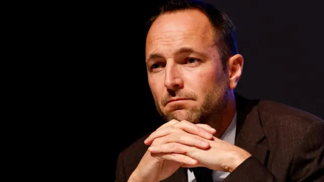 Mathias Reynard - head of the regional government of the Valais Canton - speaking at a press conference. He is using a large microphone and is wearing a dark suit and tie