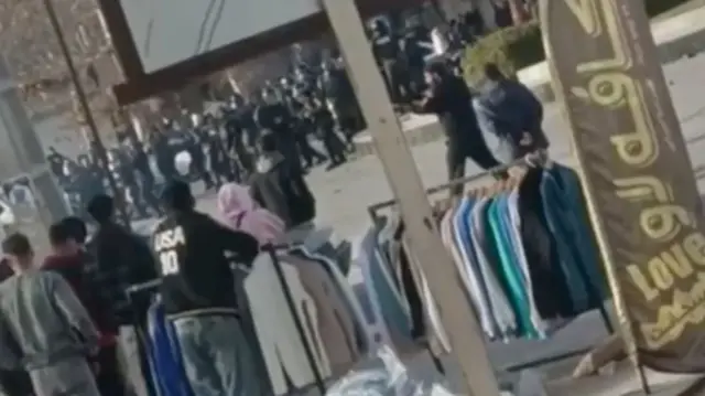 Riot police can be seen in the background of this shot, along with a man holding a gun in the middle ground and onlookers in the foreground