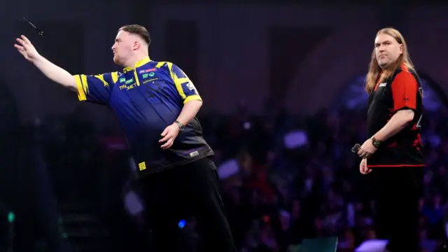 Luke Littler throws a dart as Ryan Searle looks on