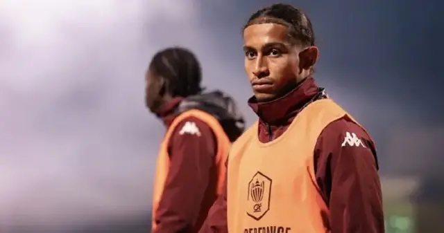 A young man in a burgundy hoodie and orange football bib stands on a sports field