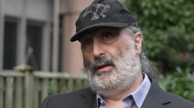 Man with a beard and wearing a cap in standing in front of a blurred building