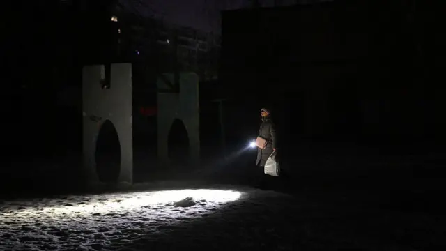 A woman walks outside in the darkness on a snow-covered street, she has light only from a torch she is carrying