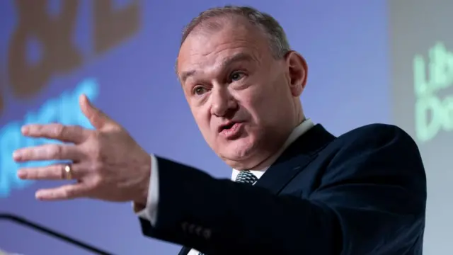Ed Davey speaking at a press conference. He's gesturing his hand outward.