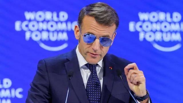 Emmanuel Macron wears blue sunglasses and a navy suit as he speaks into a mircrophone at the Davos World Economic Forum