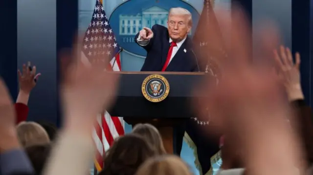 Donald Trump behind a podium pointing into a crowd of reports who have their hands raised