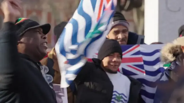 A group of people protest in central London. Some are holding the flag of the British Indian Ocean Territory.