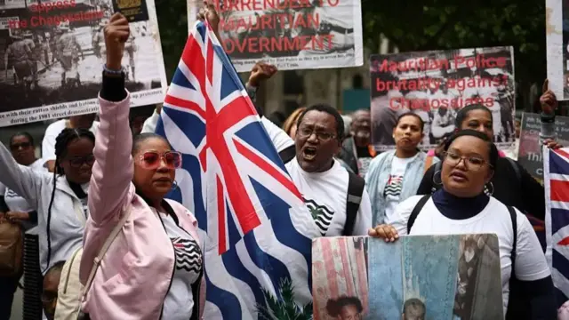 Members of the Chagossian community and supporters protest outside the High Court, holding flags and placards. One woman holds up a British passport.