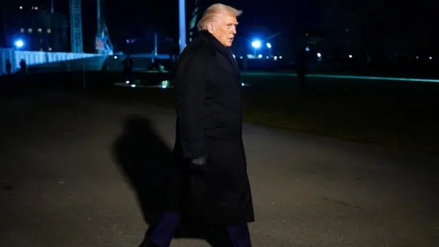 Trump p walks on the South Lawn of the White House, in the darkness, wearing a long black coat