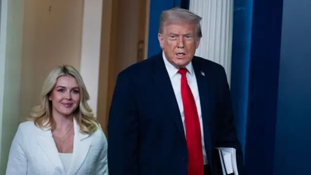 Karoline Leavitt in white next to Trump in a black suit with a red tie