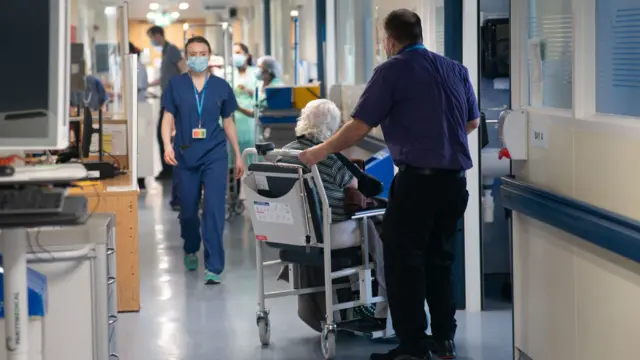 A busy hospital corridor. A young female doctor wearing blue scrubs and a face make walks towards the camera. A male orderly pushes an elderly woman in a wheelchair away from the camera. In the background is various medical paraphrenalia