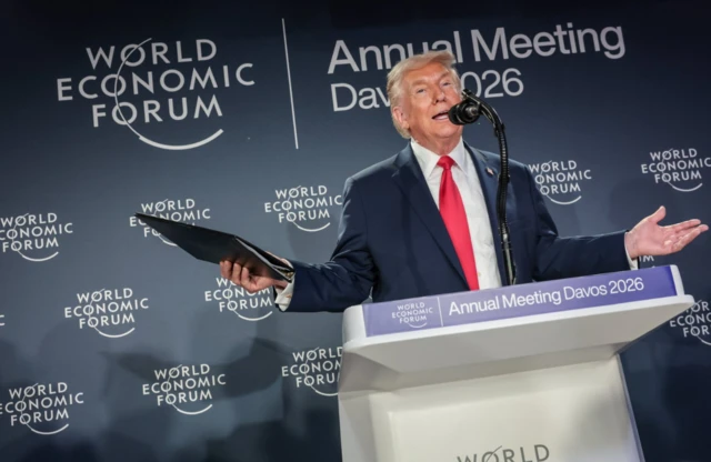 Wide-shot of Donald Trump standing behind a white lectern with the a blue sign in the top front reading "Annual Meeting Davos 2026". He's in a dark suit, white shirt and red tie, his arms open sideways, he's holding a black folder in his right hand