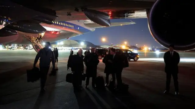 A group of people in black jackets stand under the wing of a plane
