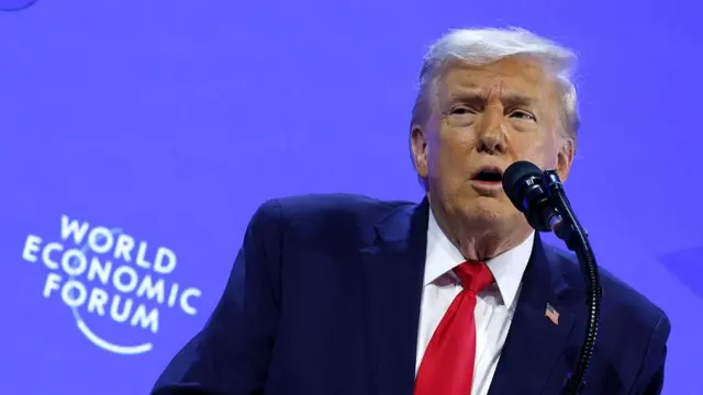 Mid-shot of Donald Trump speaking at World Economic Forum in Davos. He's standing on the right side of the frame, wearing a blue suit, white shirt and red tie - a US flag pin on his left lapel. He's speaking into a black mic, behind him a blue wall with the WEF logo in white