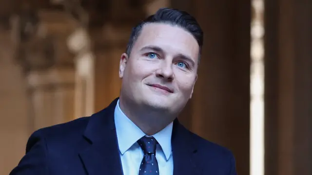 Wes Streeting smiles while looking slightly to the right of the camera. He has short brown hair and blue eyes, and is clean shaven. He is wearing a navy blue suit and tie.