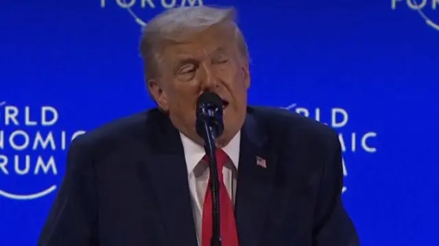 Donald Trump speaking into a microphone while standing in front of a blue background with the words 'World Economic Forum'