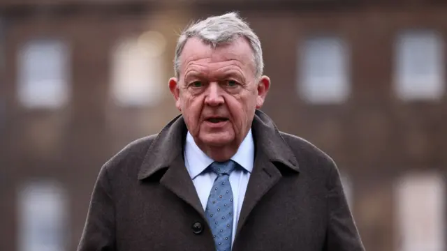 Danish foreign minister Lars Lokke Rasmussen walking towards the camera wearing a brown felt jacket on top of a light brown shirt and blue stripey tie. He looks like he is outside walking away from a building