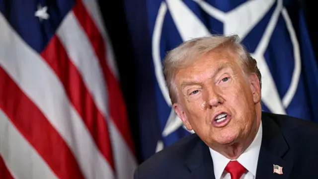Mid-shot of Donald Trump in dark suit, a US flag (L) and a Nato flag (R) behind him