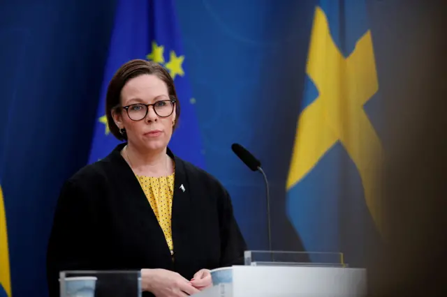 Maria Malmer Stenergard wearing a black jacket and yellow top in front of a blue background and a swedish blue and yellow flag, speaking at a podium